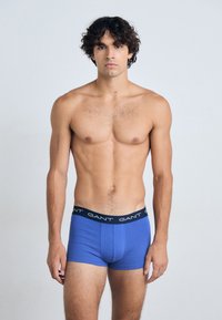 Boxers ajustados de color azul, hechos de algodón, con una cinturilla negra que presenta el nombre de la marca "GANT" en blanco. Textura suave, diseño ceñido.