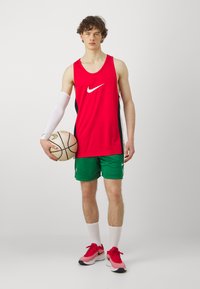 Punane Nike maika mustade külgpaneelidega, rohelised võrkpüksid, valge kokkusurumise käeslee, hoiab korvpalli, jalas punased spordijalatsid.