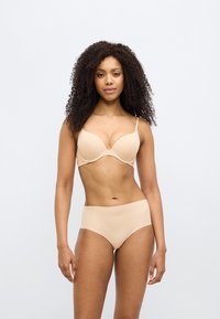 Γυναίκα με σγουρά μαλλιά που φοράει μπεζ σουτιέν και εσώρουχο ψηλόμεσο, στέκεται μπροστά σε μονόχρωμο ανοιχτό φόντο.