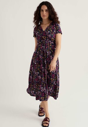 Žena s kučeravými tmavými vlasmi v námmornícky modrých kvetovaných midi šatách a bordových sandáloch stojí na svetlom jednoduchom pozadí.