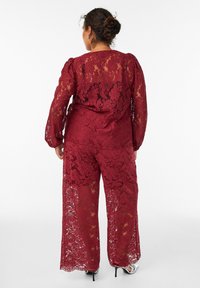 Combinaison en dentelle bourgogne avec des manches longues et un pantalon large. Détails de motifs floraux, avec une texture translucide et des bords festonnés. Talons hauts.