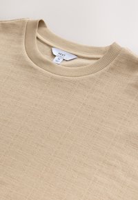 Beige crew-neck sweater made of textured fabric, featuring ribbed neckline and subtle pattern throughout. Tag shows brand "NEXT," size "M."