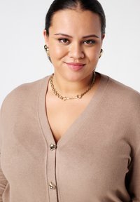 Beige cardigan met een V-hals en drie gouden knopen. Heeft een zachte textuur en wordt aangevuld met een gouden ketting en oorbellen.