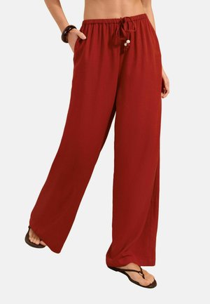 Woman wearing loose red wide-leg pants with drawstring waist and pockets, paired with black flip-flops and a black bracelet.