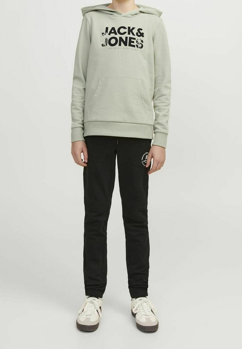Personne portant un sweat à capuche vert clair avec le texte « "JACK & JONES" », un pantalon noir avec un logo sur la cuisse, et des baskets blanches avec des semelles noires.