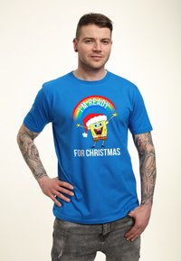 Camiseta de algodón azul con un gráfico de un personaje de dibujos animados sonriente con un gorro de Santa y el texto "ESTOY LISTO PARA NAVIDAD" en blanco.