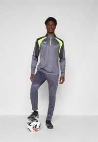 Tuta sportiva grigia con accenti giallo neon, dotata di una parte superiore con zip a quarto e pantaloni aderenti. Un pallone da calcio nero è a terra.