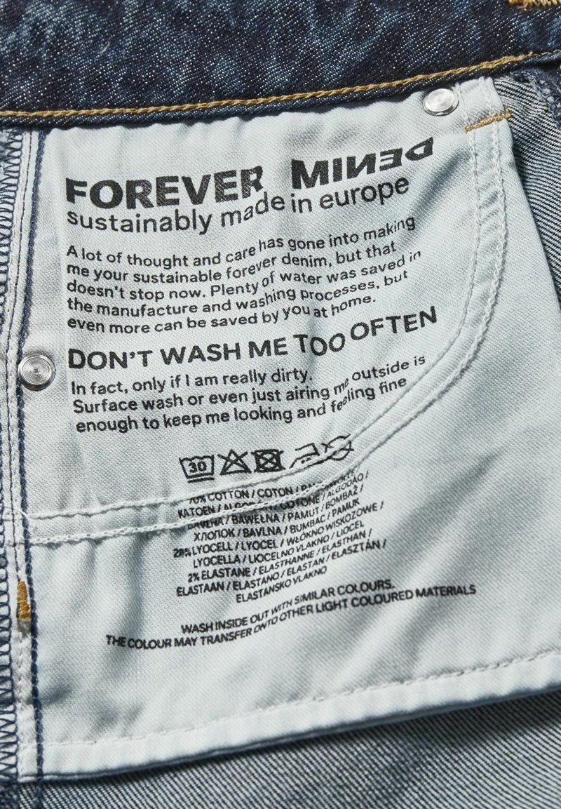 Étiquette en denim avec un tissu bleu foncé. Le texte comprend "FOREVER MINED", des instructions d'entretien et des détails sur la composition du tissu en plusieurs langues.