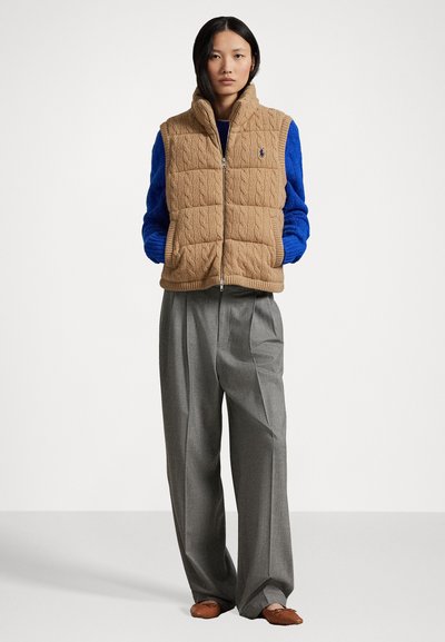 Gaišpelēka quilted veste ar augstu apkakli un adījuma tekstūru, uzvilkta virs zilā džempera un saskaņota ar pelēkām krokotām biksēm.