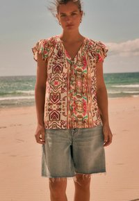Blouse à manches courtes à motifs en crème avec des motifs géométriques roses, rouges et verts ; associée à un short en denim bleu clair avec un ourlet effiloché.