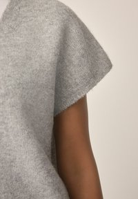 Pull en tricot gris doux à manches courtes porté sur un bras avec un teint de peau moyen foncé sur un fond neutre.