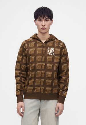 Junger Mann mit kurzen schwarzen Haaren trägt einen braunen karierten Zip-Hoodie mit weißem Motiv auf der Brust und hellgraue Hosen.