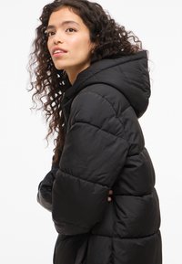 Crna dukserica s kapuljačom i prošivenim dizajnom, s opuštenim krojem i mekanim, sjajnim tkaninom. Rukavi su naborani i izolirani.