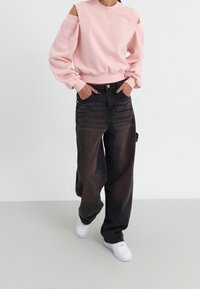 Light pink sweatshirt with cut-out shoulders, long sleeves, and ribbed hem; paired with loose black denim pants and white sneakers.