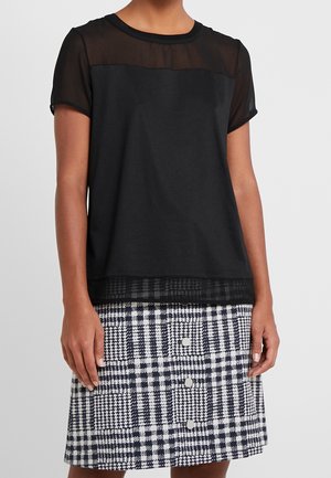 Mujer que lleva una camiseta negra de manga corta con panel superior transparente y una falda de cuadros en blanco y negro con tres botones delanteros.