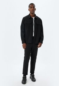 Zwarte button-up jas met twee borstzakken, gedragen over een witte shirt. Gepaard met zwarte slim-fit broek en zwarte sneakers. Minimalistisch ontwerp.