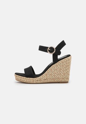 Sandalia de cuña negra con tiras de lona, hebilla circular y tacón trenzado de yute. La suela es marrón con una textura suave.