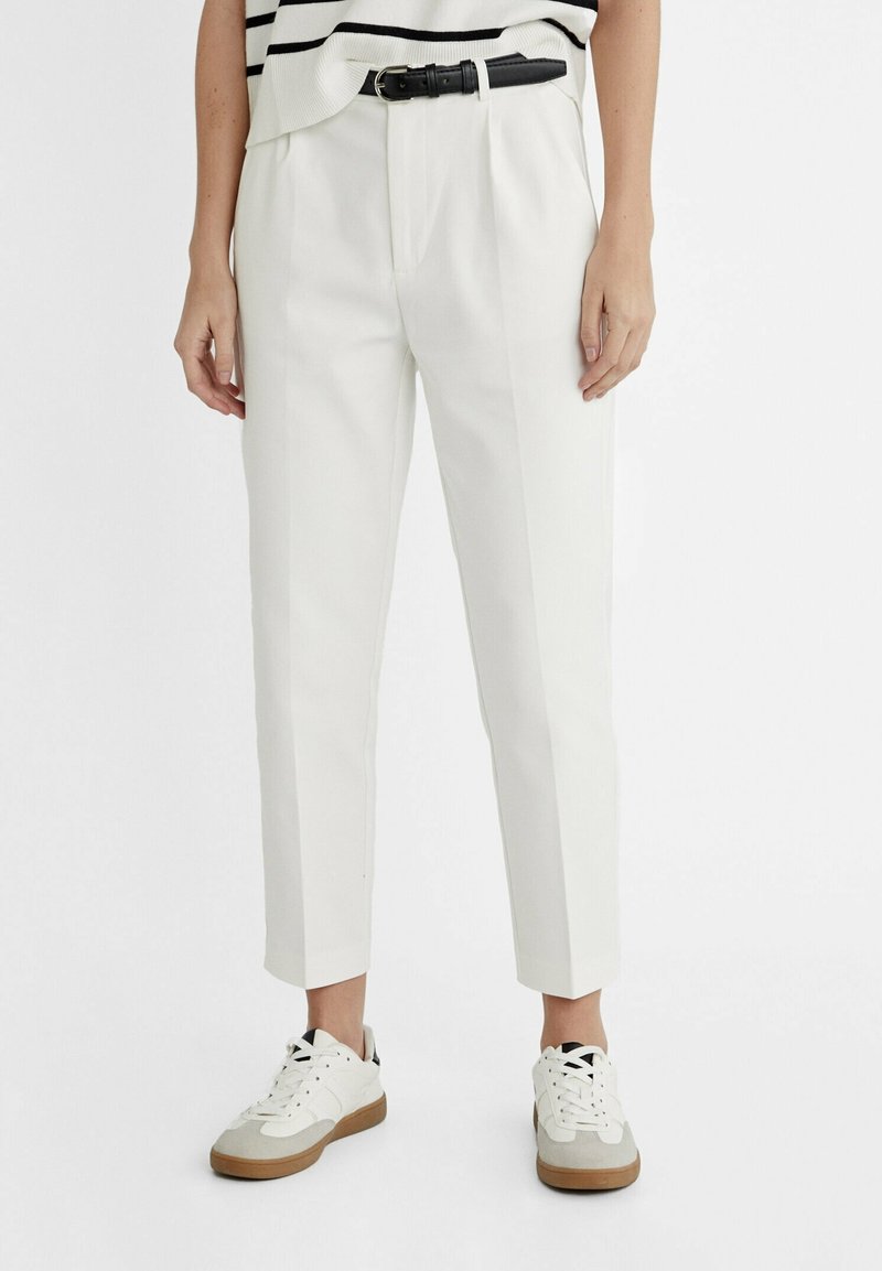 Witte op maat gemaakte broeken met een hoge taille, voorzien van een zwarte riem en zijzakken. Ze worden gecombineerd met casual witte sneakers.