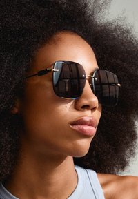 Fekete négyzet alakú napszemüveg arany fém kerettel és díszítő részletekkel. A sima lencsék visszaverik a fényt. A modellen göndör haj és semleges bőrszín látható.