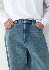 Hellblaue Jeans aus Denim mit hoher Taille, fünf Taschen und kontrastierender Naht. Ein silberner Knopf und kupferfarbene Nieten sind sichtbar.