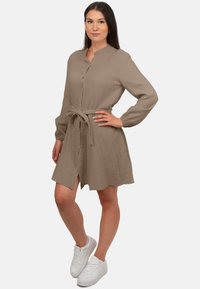 Abito beige con bottoni, cintura in vita, maniche lunghe e tessuto testurizzato. Indossato con sneaker bianche, caratterizzato da una fessura frontale.