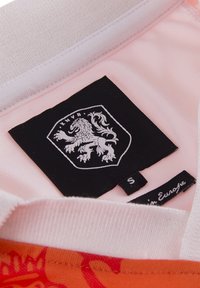 Kraag van een kortärmige kledingstuk in witte stof met een roze binnenvoering, voorzien van een zwart label met een leeuwembleem en een maatlabel.