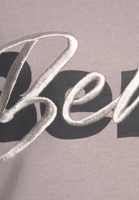 Auf einem weichen, hellrosa Stoff gestickter Text: ein glänzendes silbernes "Sei" neben fettem, mattem schwarzen Schriftzug, der einen strukturierten Kontrast schafft.