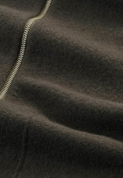 Dunkelgrauer Wollstoff mit weicher Textur und sichtbaren Nähten. Ein goldfarbener Metallreißverschluss verläuft entlang des Stoffes und betont Tiefe und Falten.