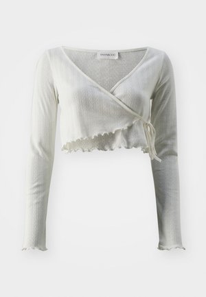 Fehér, rövidített, hosszúujjú felső V-dekoltázzsal, bordázott textúrával és fodros szegéllyel. Összehúzott dizájnú, deréknál megkötős.