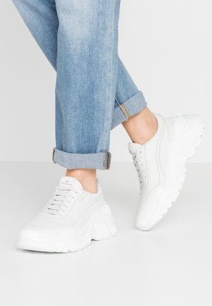 Baskets blanches épaisses avec lacets portées avec un jean bleu clair retroussé, montrant les chevilles sur un fond gris clair uni.