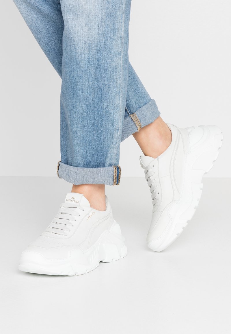 Baskets blanches épaisses avec lacets portées avec un jean bleu clair retroussé, montrant les chevilles sur un fond gris clair uni.