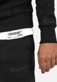 Felpa nera con polsini a coste e logo ricamato. La vita bianca presenta un'etichetta evidente. La trama del tessuto appare morbida e confortevole.