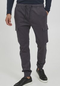 Cargo broek in donkergrijs katoen, met een trekkoord in de taille, elastische enkelboorden en twee zijzakken met kleppen. Zwarte sneakers.