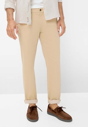 Mand iført beige chinos med op-rullede manchetter, brune snøresko, hvid skjorte stoppet ind under en beige knappet skjorte.