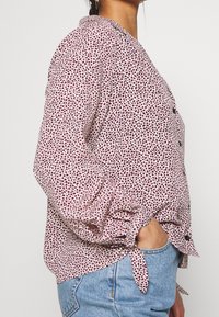 Knappad blus i ljus krämfärg med röda och svarta prickar. Har krage, långa ärmar med samlade manschetter och knytdetalj i midjan.