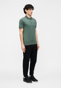 Polo verde com um pequeno logotipo bordado, combinado com calças de treino pretas. As calças têm um detalhe dourado na lateral. Ténis pretos completam o visual.