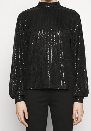 Top noir à manches longues avec sequins, col montant et manches à poignets porté par une personne visible du cou jusqu'aux hanches sur un fond uni.
