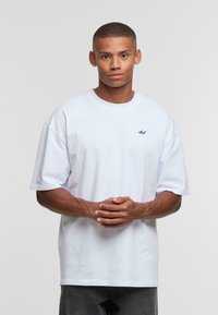 Camiseta de algodón blanca con mangas cortas, escote redondo y pequeño logo azul marino en la parte izquierda del pecho. Combinada con pantalones grises.