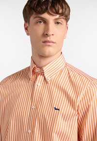 Kortärmad skjorta med knappar som har orange och vita vertikala ränder, en klassisk krage och en liten blå broderad logotyp på bröstet.