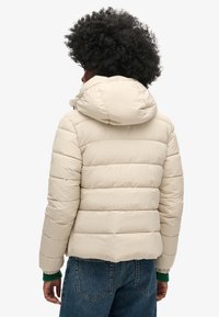 Beige vadderad jacka med huva, med horisontell quiltning, elastiska ärmslut och avslappnad passform. Synliga sömmar och slät textur.