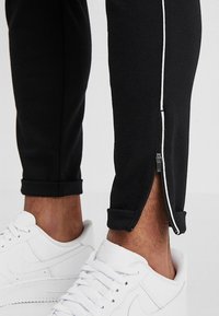 Schwarze sportliche Jogginghosen mit weißen Seitenreißverschlüssen und Paspelierung, mit einem Bündchen am Saum. Kombiniert mit weißen Sportschuhen.