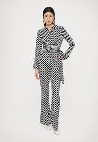 Melnā un baltā krāsā ar rakstu wrap stila kombinezons ar garām piedurknēm un plašām biksēm, ar sasienamu jostu un ģeometriskiem motīviem.