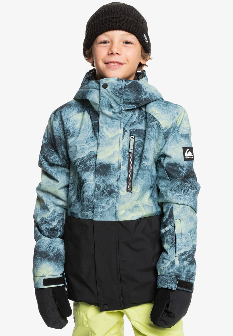 Mönstrad snöjacka i blått och svart, med en dragkedjeförsedd bröstficka, justerbara ärmmuddar och huva, bärs tillsammans med svarta handskar och ljusa byxor.