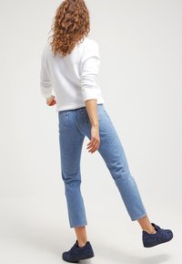 Vit sweatshirt, ljusblå cropped jeans och mörkblå sneakers. Lockigt hår och hand på höften, som visar en avslappnad outfit från sidan.