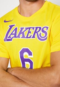Hombre con camiseta amarilla de los Lakers con el número 6 en púrpura, brazos cruzados sobre el pecho, vista cercana del torso y cuello.