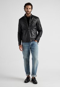 Schwarze Lederjacke mit gesteppten Schulterdetails, vorderen Reißverschlüssen, zwei Seitentaschen, kombiniert mit hellblauen, umgeschlagenen Jeans und schwarzen Slippern.
