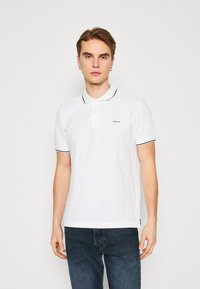 Witte poloshirt met zwarte kraagaccenten. Heeft korte mouwen en een knoopsluiting. Logo weergegeven op de linkerkant van de borst. Gladde textuur.