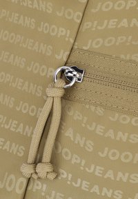 Beżowa torba materiałowa z powtarzającym się logo "JOOP!JEANS". Wyposażona w srebrny zamek błyskawiczny z okrągłym uchwytem i węzłem z materiału.