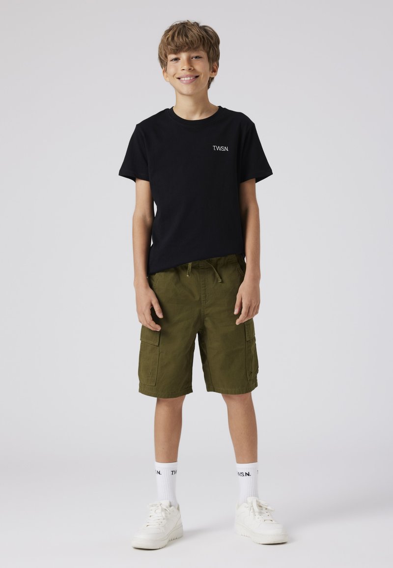 T-shirt nera in cotone con logo "TWSN", pantaloni cargo verdi con tasche laterali, calzini bianchi e sneakers bianche su uno sfondo neutro.