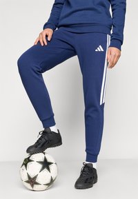 Σπορ παντελόνι ναυτικού μπλε με λευκές ρίγες, ριμπ μανσέτες και πλαϊνές τσέπες, σε συνδυασμό με μαύρα αθλητικά παπούτσια σε ποδοσφαιρική μπάλα.